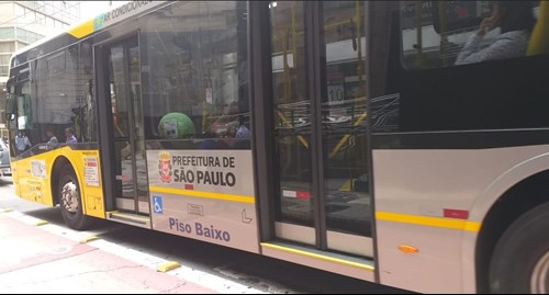 #PraCegoVer: Foto da lateral de um ônibus articulado amarelo, se locomovendo pelo centro da cidade