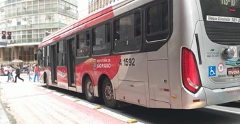 #PraCegoVer: Imagem de um trólebus de cor vermelha e cinza, que atende ao centro e a Zona Leste. O veículo está parado no semáforo, enquanto os pedestres atravessam a rua.