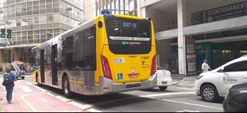 #pracegover traseira do ônibus amarelo área três perto de carros brancos e preto ao lado ciclofaixa