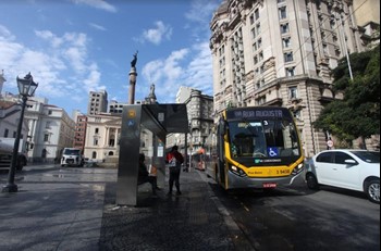 #pracegover ônibus amarelo perto do ponto ao lado de carro branco