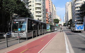 #pracegover três ônibus um da cor prata um da cor verde e um azul em sentidos diferentes com prédios e ciclofaixas na avenida