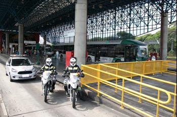 #PraCegoVer: Imagem de dois funcionários da SPTrans em motos, com um carro da empresa atrás e um ônibus de cor verde ao fundo, no Terminal Mercado